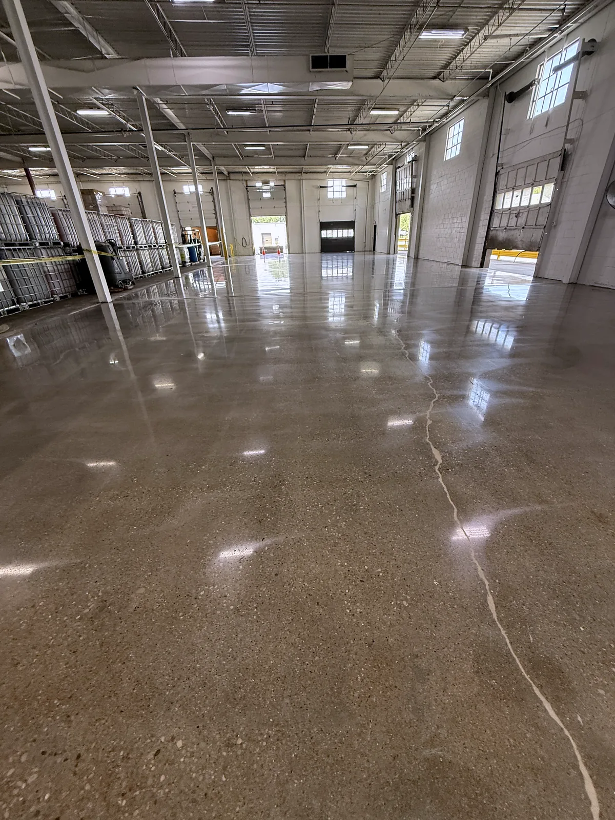 Long industrial bay with dock doors in the distance, polished concrete floor with a hairline crack in the foreground, Pittsburgh area.