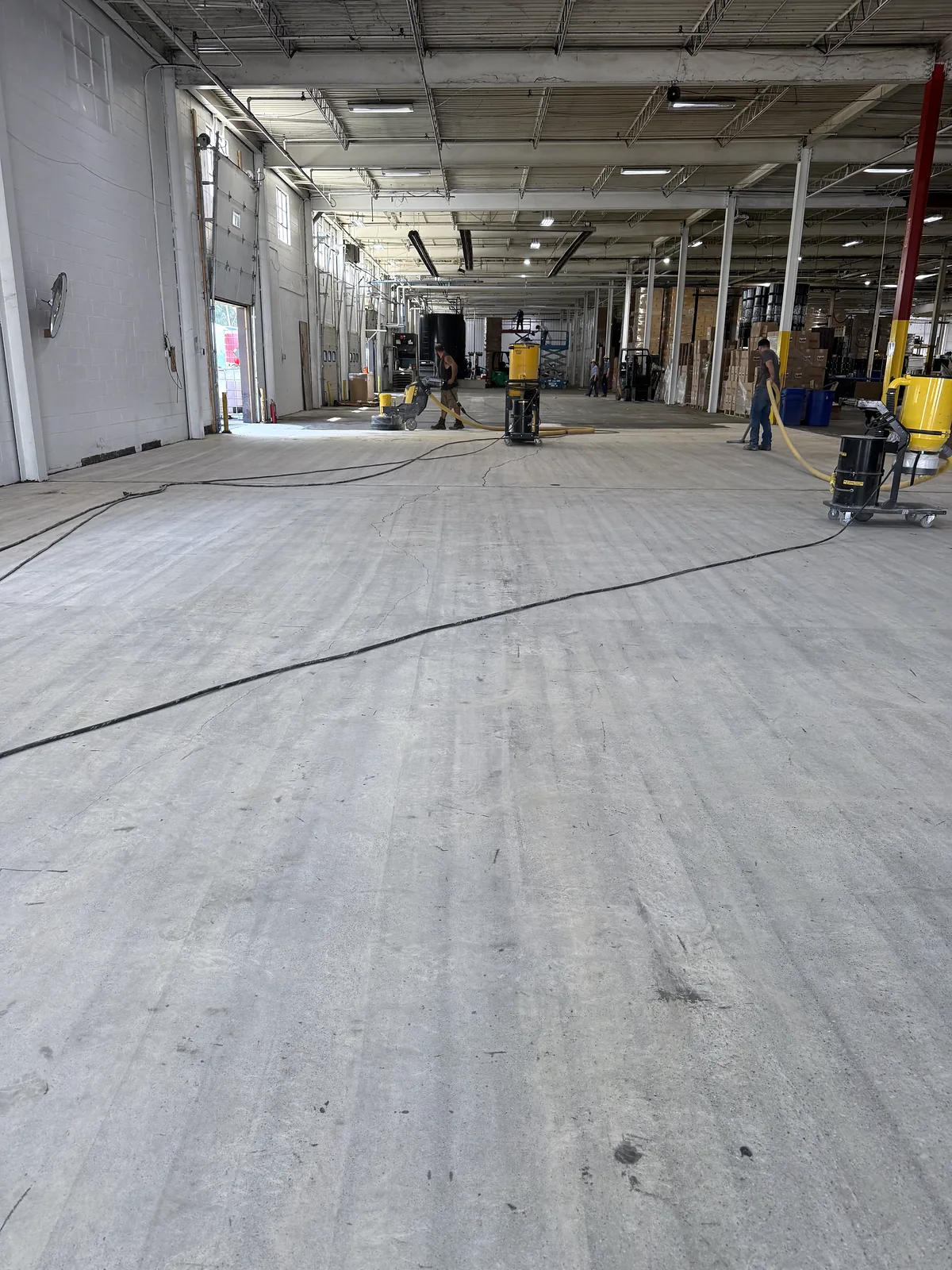 Large warehouse floor being ground, workers and dust extractors in the distance, cord across the floor, Pittsburgh area.
