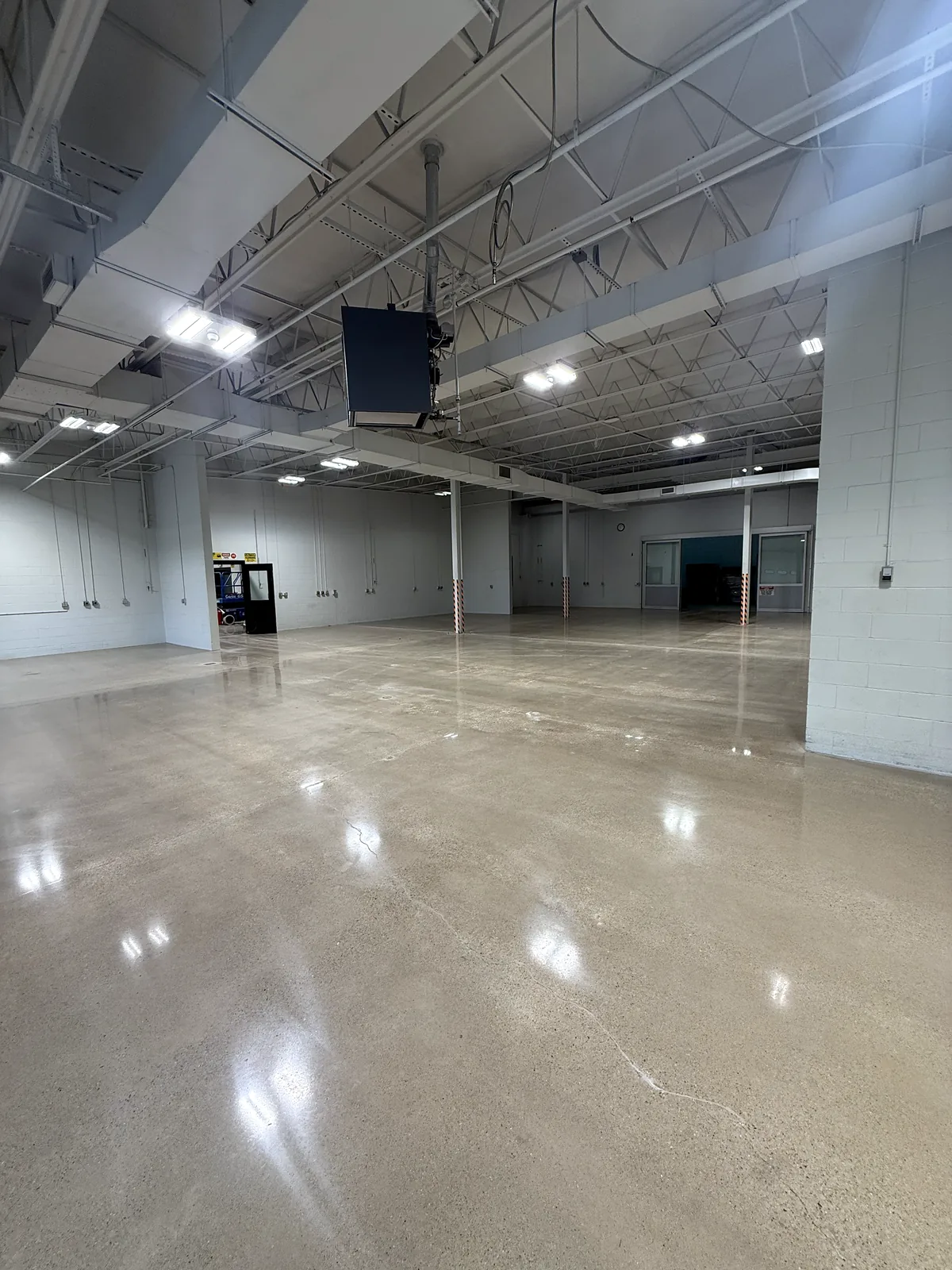 Wide empty warehouse bay with a hanging black unit, polished concrete floor reflecting ceiling lights, one fine hairline crack visible, Pittsburgh area.